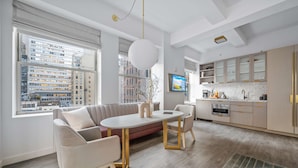 A modern living room with a sofa, dining table, and chairs, a kitchen with cabinets and appliances, and large windows offering city views. - 17john: Deluxe King Studio Apartment (New York)