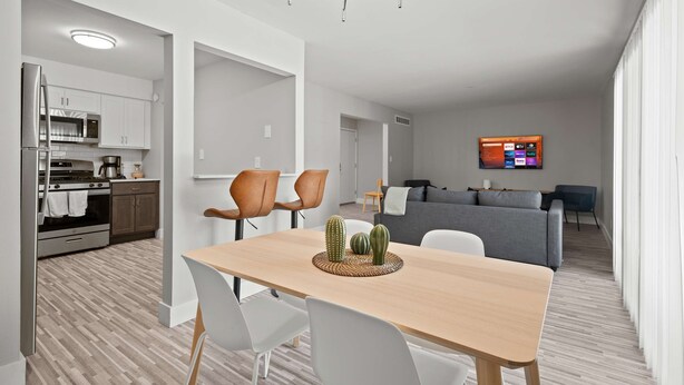 Eine moderne Küche mit weißen Schränken, einem eingebauten Ofen und einer Mikrowelle. Es gibt einen Essbereich mit einem Holztisch und weißen Stühlen. Im Hintergrund ist ein Wohnzimmer mit einem grauen Sofa und einem an der Wand befestigten Fernseher zu sehen.