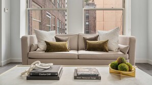 Un salon moderne avec un canapé beige, deux fenêtres donnant sur la ville et une table basse agrémentée de livres et de boules décoratives.