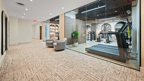 Un intérieur moderne avec un sol recouvert de moquette, un coin salon avec des fauteuils et un mur de verre donnant sur une salle de sport équipée de matériel d'exercice.