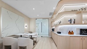 Une chambre d’hôtel moderne au design minimaliste, comprenant un comptoir en bois aux lignes courbes, un coin salon avec une table et des chaises, ainsi qu’une grande œuvre d’art abstraite ornant le mur.