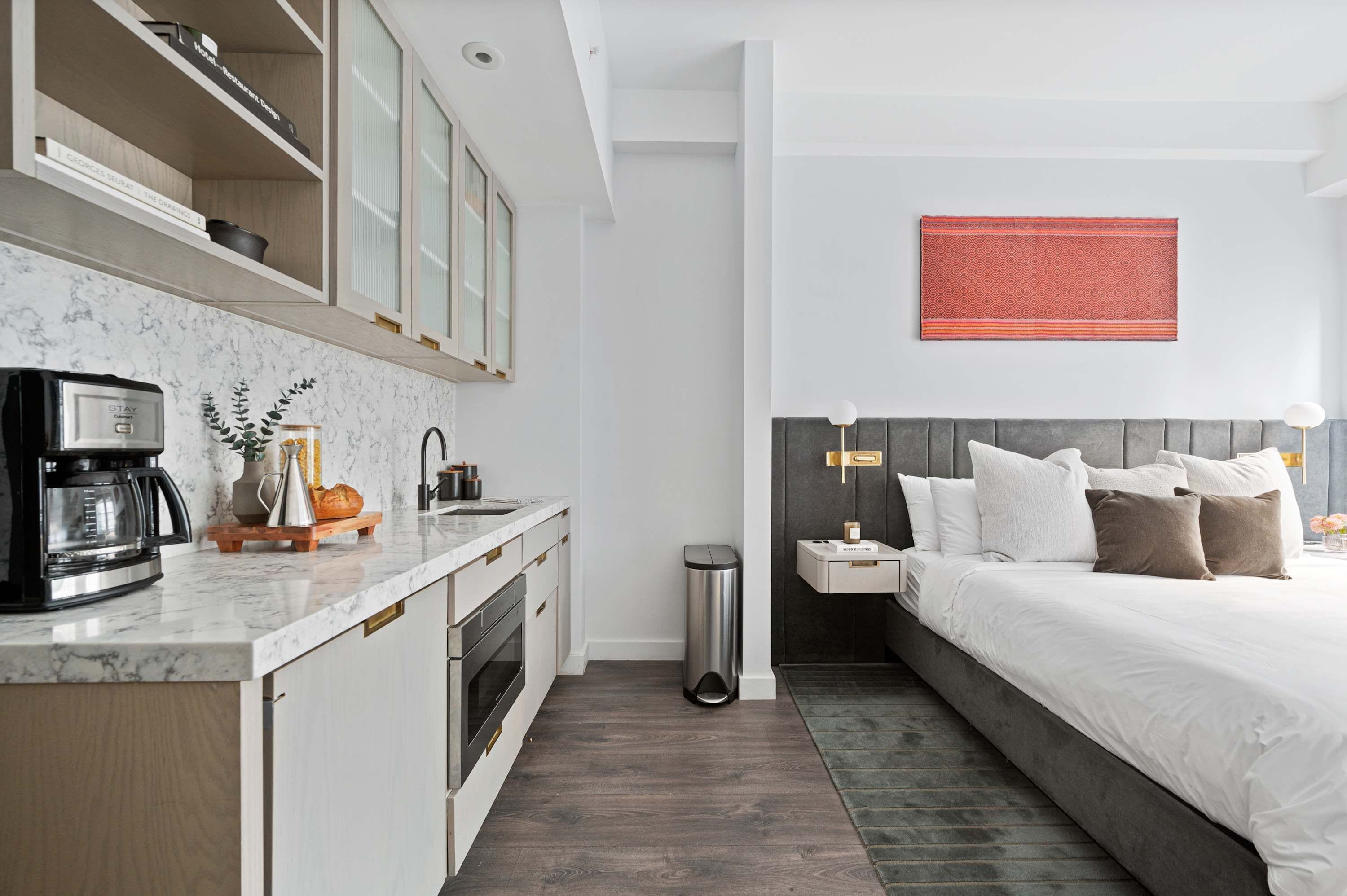 A modern hotel room with a kitchen area, a bed with a grey headboard, and a red patterned wall hanging.