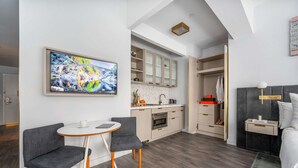 A modern hotel room with a dining area, kitchenette, and a large TV mounted on the wall.