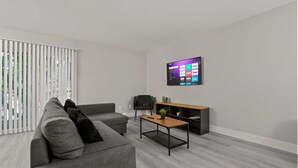 A modern living room with a grey sofa, a coffee table, a TV mounted on the wall, and a window with blinds. - Cozy 2BR 2BA Apartment for the Family (Rochester)