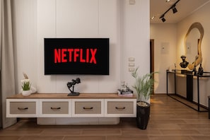 Un soggiorno moderno con una TV a schermo piatto che mostra 'NETFLIX', un tavolino consolle in legno con oggetti decorativi, una pianta in vaso e uno specchio a parete.