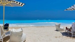A beach with white lounge chairs and striped umbrellas, a clear blue sky, and the ocean in the background.