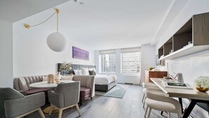A modern hotel room with a sofa, dining table, chairs, a bed, and a view of the city through large windows. - 17john: Junior King Studio (New York)