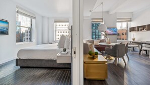 A modern hotel room with a large bed, a desk, a dining area, and a view of the cityscape.
