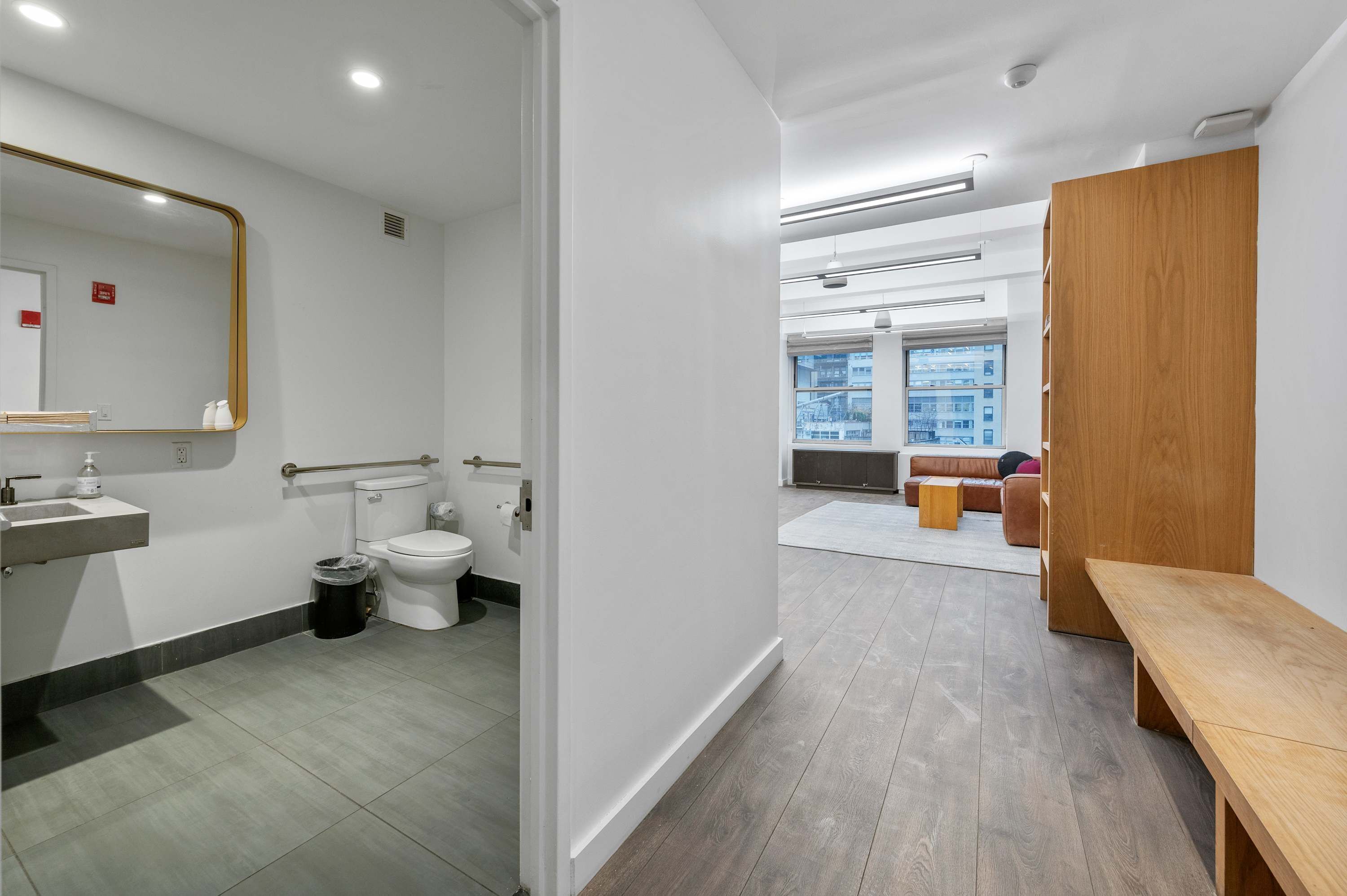 A modern bathroom with a toilet, sink, and mirror. There is a wooden bench and a view of an outdoor area with a building.