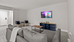 En moderne stue med en grå sofa, to lænestole, et sofabord og et vægmonteret fjernsyn, der viser streamingtjenester.