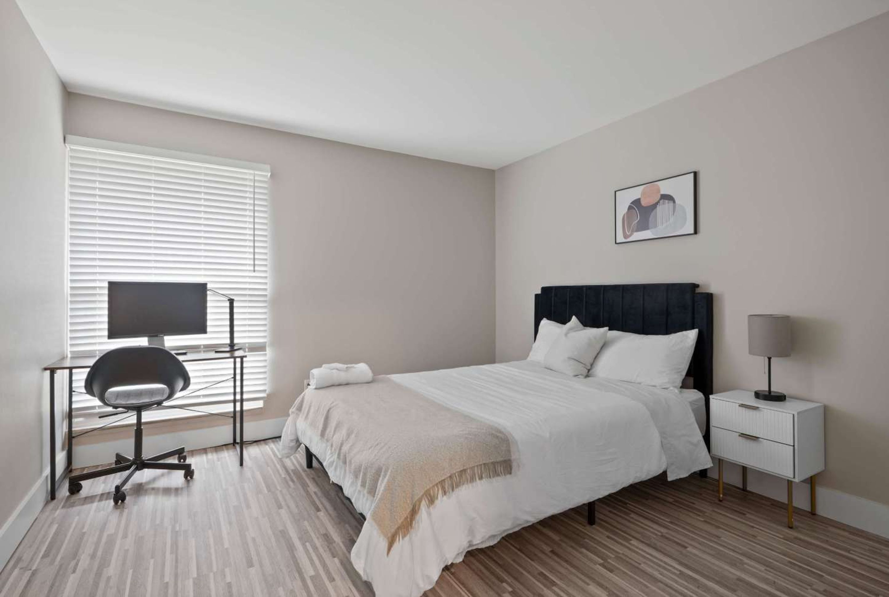A bedroom with a bed, a desk with a chair, a TV, a bedside table with a lamp, and a framed picture on the wall.