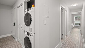 A laundry room with stacked washers and dryers, a wooden floor, and a hallway leading to a bedroom with a window. - Tranquil 2BR 2BA Getaway for the Family (Rochester)