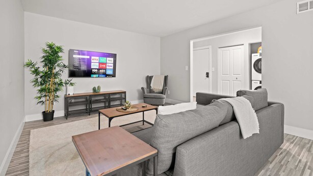 A modern living room with a grey sofa, a coffee table, a TV mounted on the wall, a potted plant, and a door leading to a laundry area. - Tranquil 2BR 2BA Getaway for the Family (Rochester)