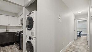 A laundry room with stacked washers and dryers, a kitchen with dark cabinets and a stainless steel range, and a hallway with a wooden floor. - Beautiful 2BR 2BA Apartment With Modern Design (Rochester)