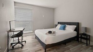 A bedroom with a bed, a desk, a chair, a nightstand, and a window with blinds. - Beautiful 2BR 2BA Apartment With Modern Design (Rochester)