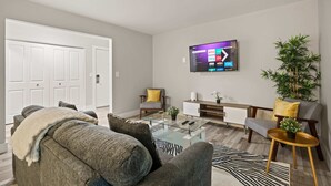 A modern living room with a grey sofa, a glass coffee table, a TV mounted on the wall, and a plant on a side table. - Beautiful 2BR 2BA Apartment With Modern Design (Rochester)