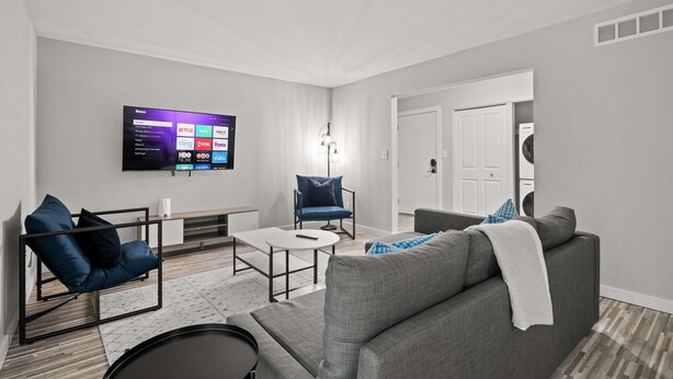 A modern living room with a flat-screen TV mounted on the wall, a grey sofa, a blue armchair, a white coffee table, and a small rug. - Family Friendly 2BR 2BA in the City (Rochester)