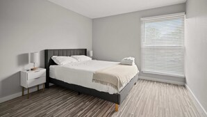 A bedroom with a bed, nightstands, a lamp, and a window with blinds. - Family Friendly 2BR 2BA in the City (Rochester)