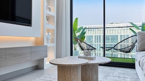 Ein modernes Wohnzimmer mit großem Fernseher, einem runden Couchtisch und Blick auf ein Gebäude durch bodentiefe Fenster.
