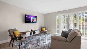 A modern living room with a flat-screen TV mounted on the wall, a grey sofa, a wooden armchair, a glass coffee table, and a patterned rug. - Bright and Airy 2BR 2BA in the Heart of the City (Rochester)