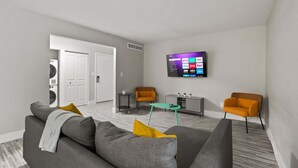 A modern living room with a grey sofa, two orange armchairs, a small teal coffee table, and a flat-screen TV mounted on the wall. - Comfy 2BR 2BA Unit in Pime Location (Rochester)