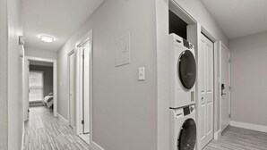 A laundry room with a front-loading washer and dryer, a hallway with doors, and a patterned floor. - Comfy 2BR 2BA Unit in Pime Location (Rochester)