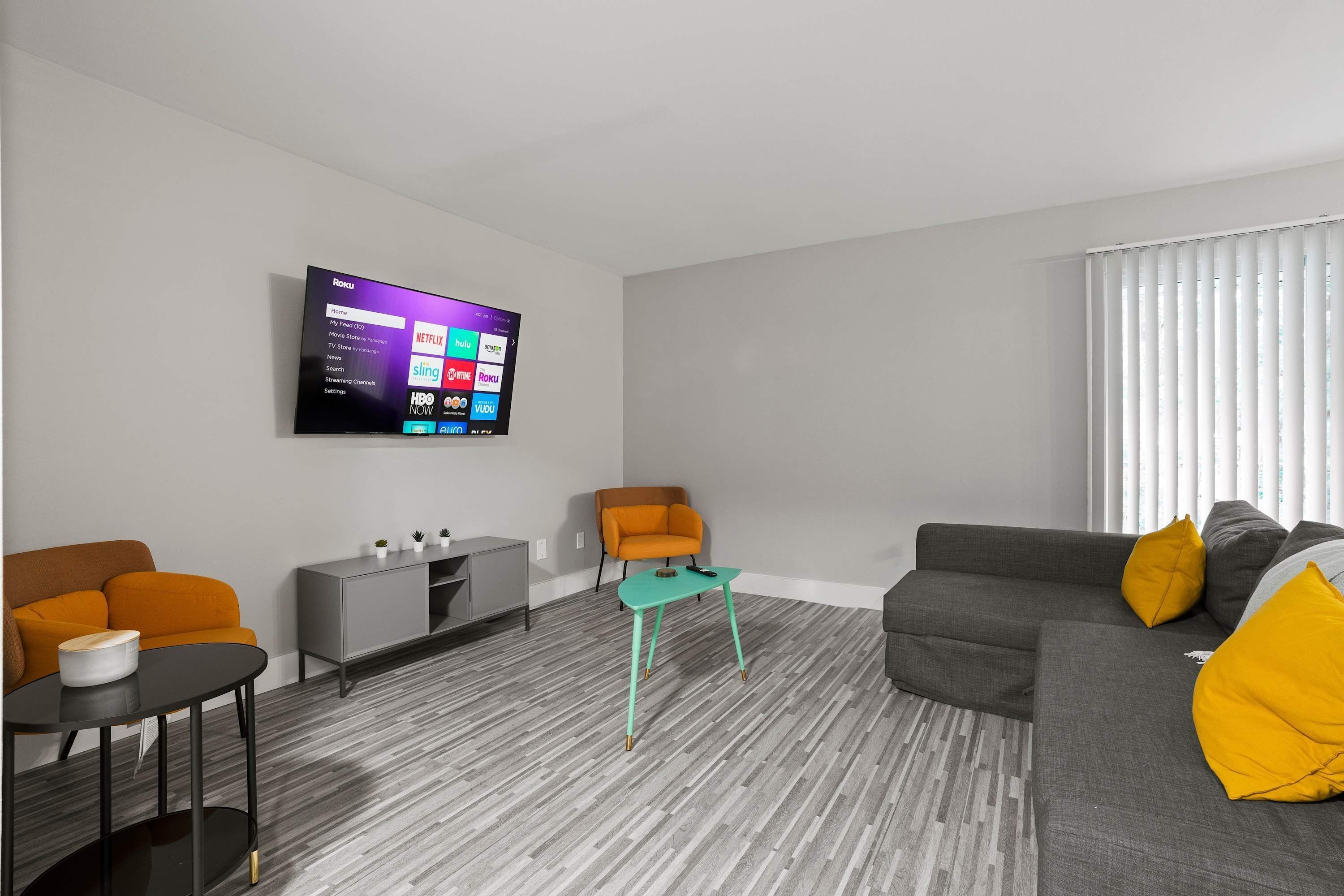 A modern living room with a flat-screen TV mounted on the wall, a grey sofa with yellow cushions, a small orange armchair, a grey cabinet, and a teal coffee table.