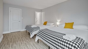 Une chambre d’hôtel avec trois lits, chacun recouvert d’une courtepointe à motifs et agrémenté d’une serviette blanche. Il y a une porte sur la gauche et une source de lumière sur la droite.