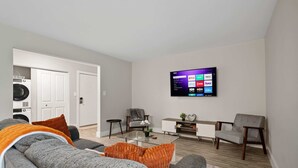 Un salon moderne avec un téléviseur à écran plat fixé au mur, des canapés gris, une table basse en verre et un meuble blanc.