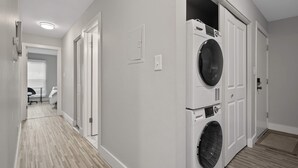 Une buanderie équipée d’un lave-linge et d’un sèche-linge à chargement frontal, une porte donnant sur un couloir et un petit coin salon avec une chaise et une table.
