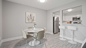 A modern kitchen with white cabinets, a stainless steel refrigerator, and a microwave. There is a dining area with a round table and chairs, and a bar area with two stools. - 2-bedroom Spacious Stay (Rochester)