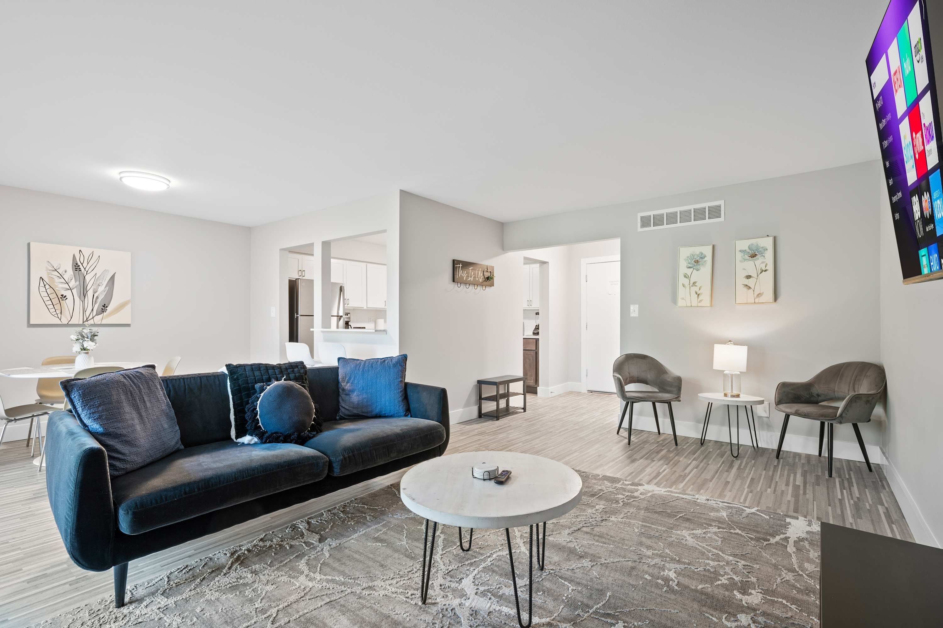 A modern living room with a black sofa, a round coffee table, and two armchairs. There is a kitchen visible in the background. - 2-bedroom Spacious Stay (Rochester)