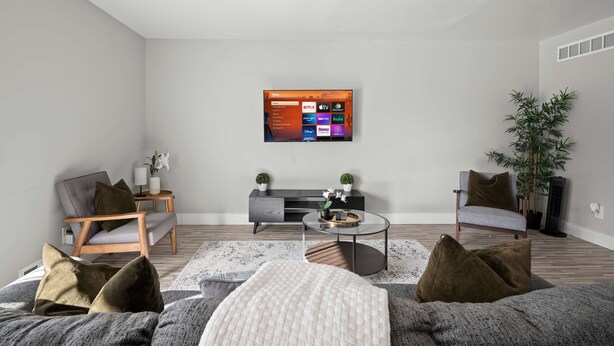 Ett modernt vardagsrum med en platt-TV som är monterad på väggen, en grå soffa med kuddar, ett litet runt soffbord och ett sidobord med en lampa och en växt.