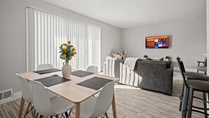 Ett modernt vardagsrum med en matplats, en grå soffa, en tv som är monterad på väggen och en växt på bordet.