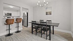 A modern kitchen with white cabinets, a stainless steel refrigerator, and a microwave. There is a dining table with four chairs and a chandelier above.
