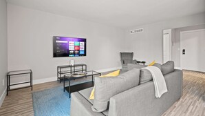 A modern living room with a flat-screen TV mounted on the wall, a grey sofa, a small coffee table, and a glass-top coffee table.