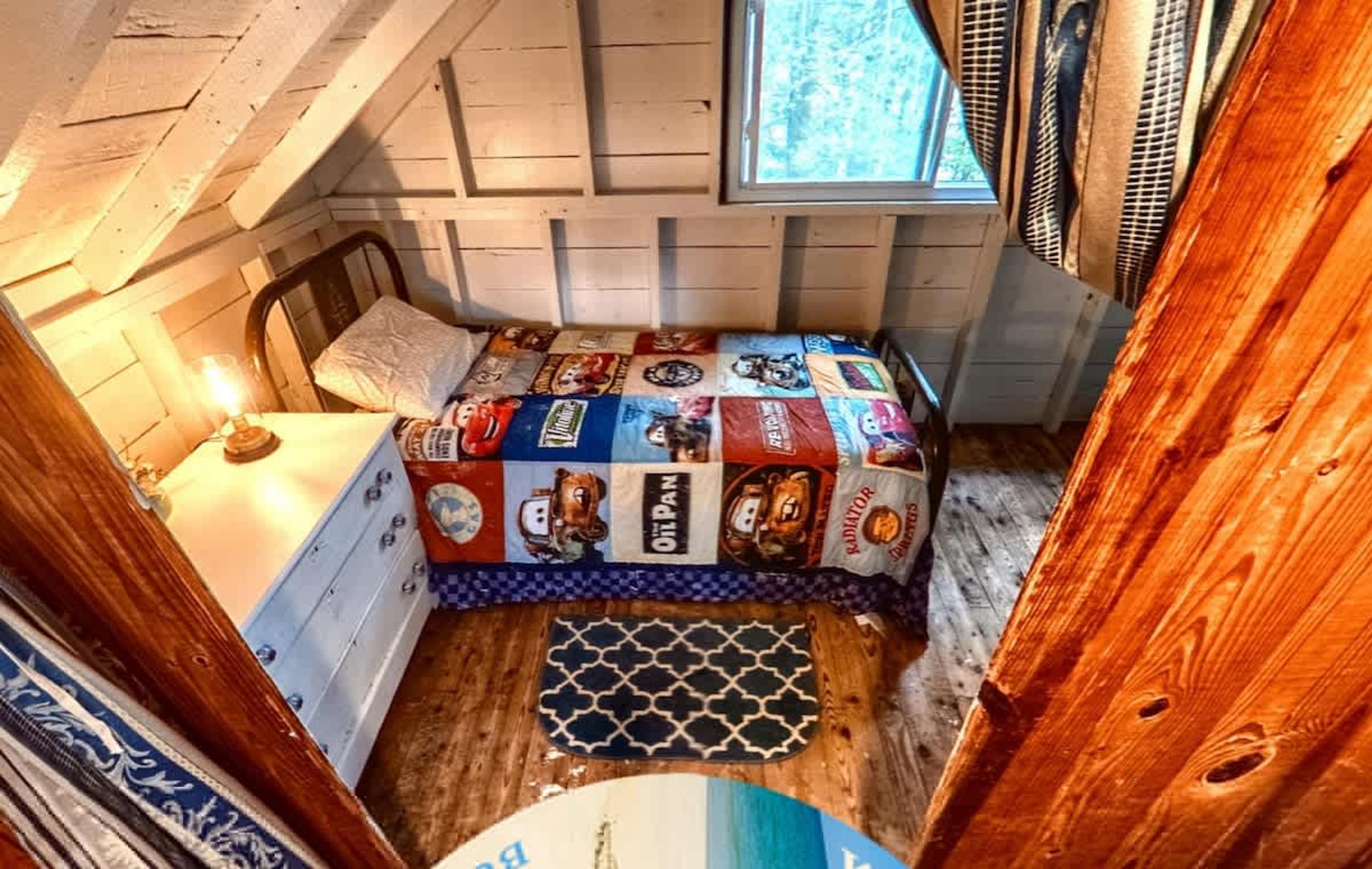 A small, cozy room with a bed covered in a patchwork quilt, a wooden dresser, a small table with a lamp, and a window with curtains.