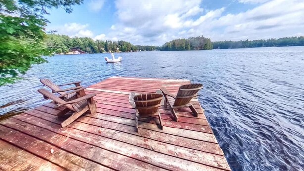 Eine Holzsteg mit zwei Adirondack-Stühlen und einer Holzschüssel, mit Blick auf einen stillen See, in der Ferne ein Segelboot und ein bewaldeter Uferstreifen.