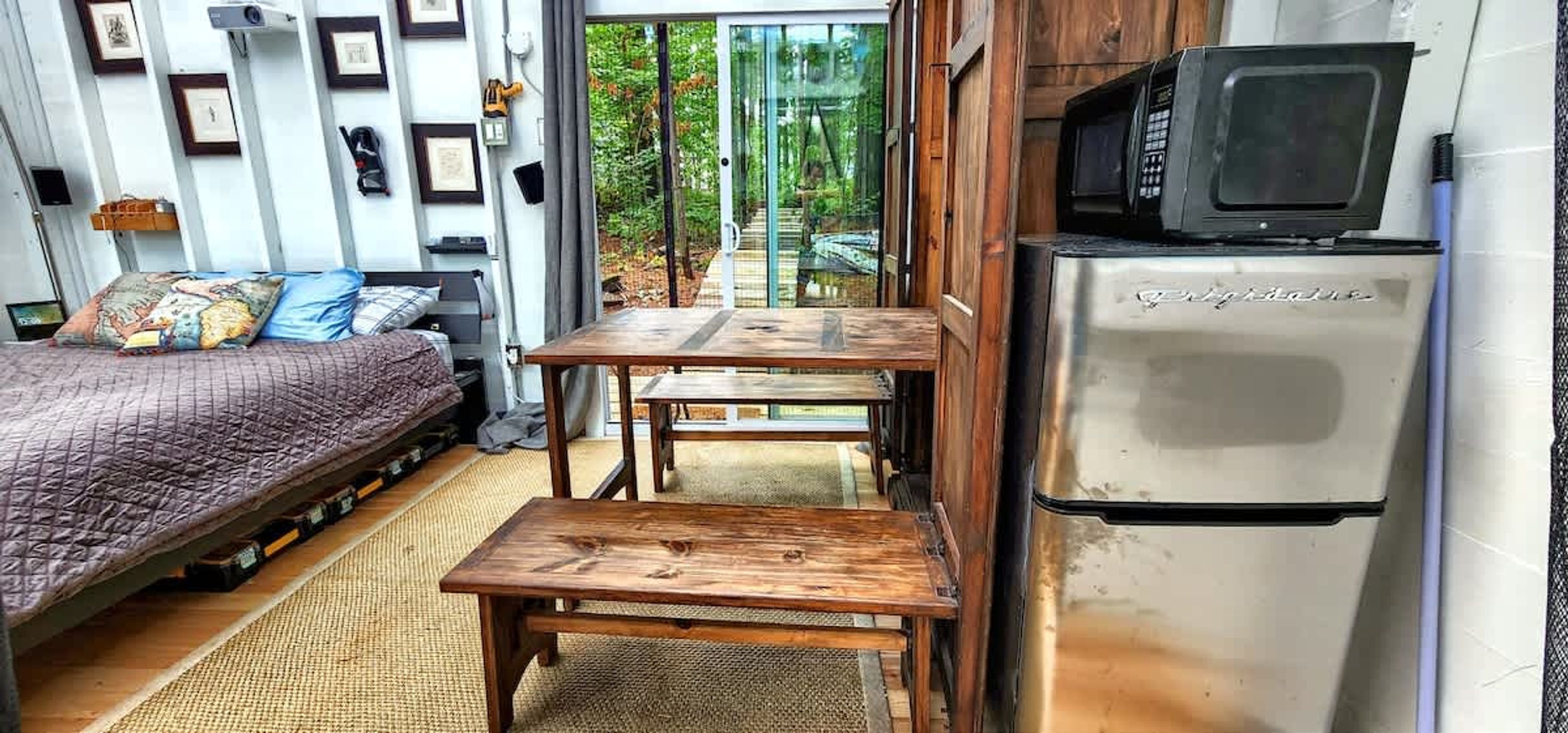 A compact room with a bed, two wooden tables, a refrigerator, and a sliding glass door leading to a garden.