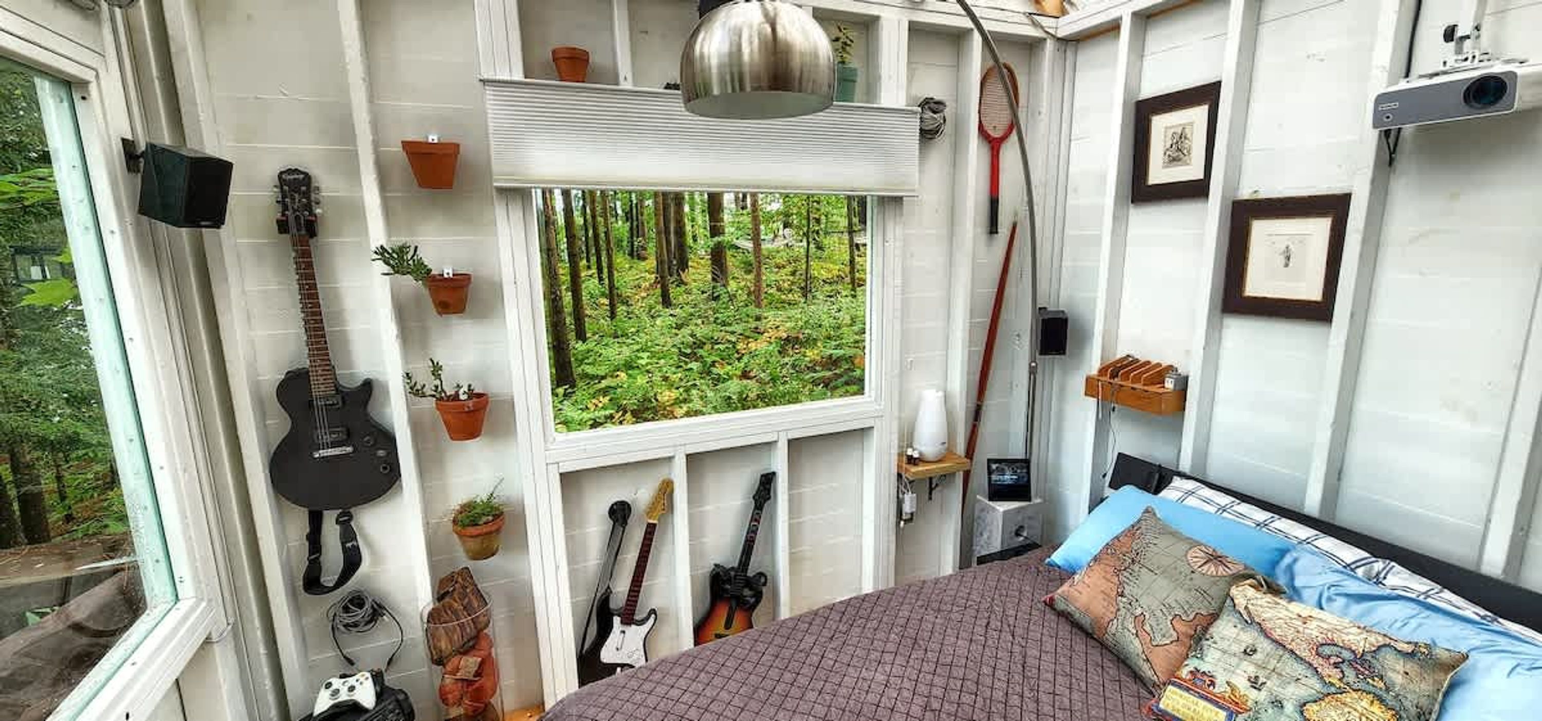 Un dormitorio con una cama, guitarras, una ventana con vista a un bosque y un proyector.