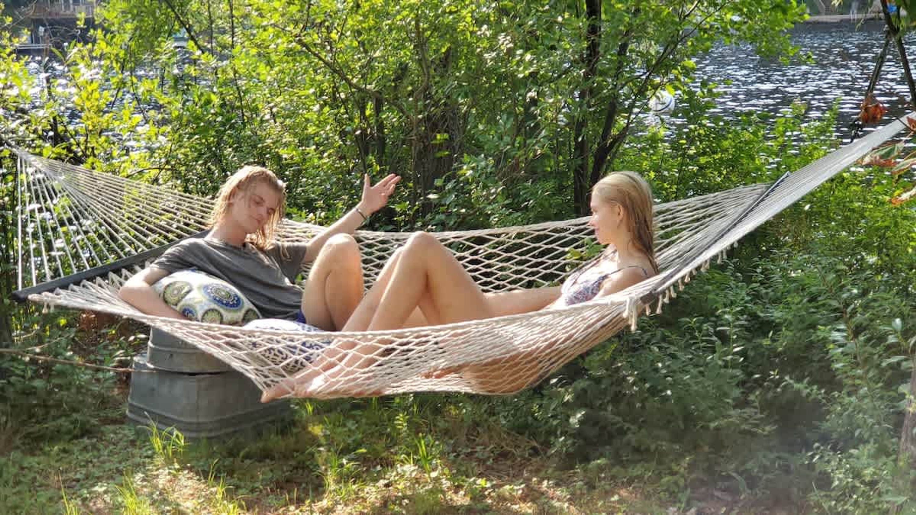 Due persone si rilassano in un'amaca all'aperto, con vegetazione e un corpo d'acqua sullo sfondo.
