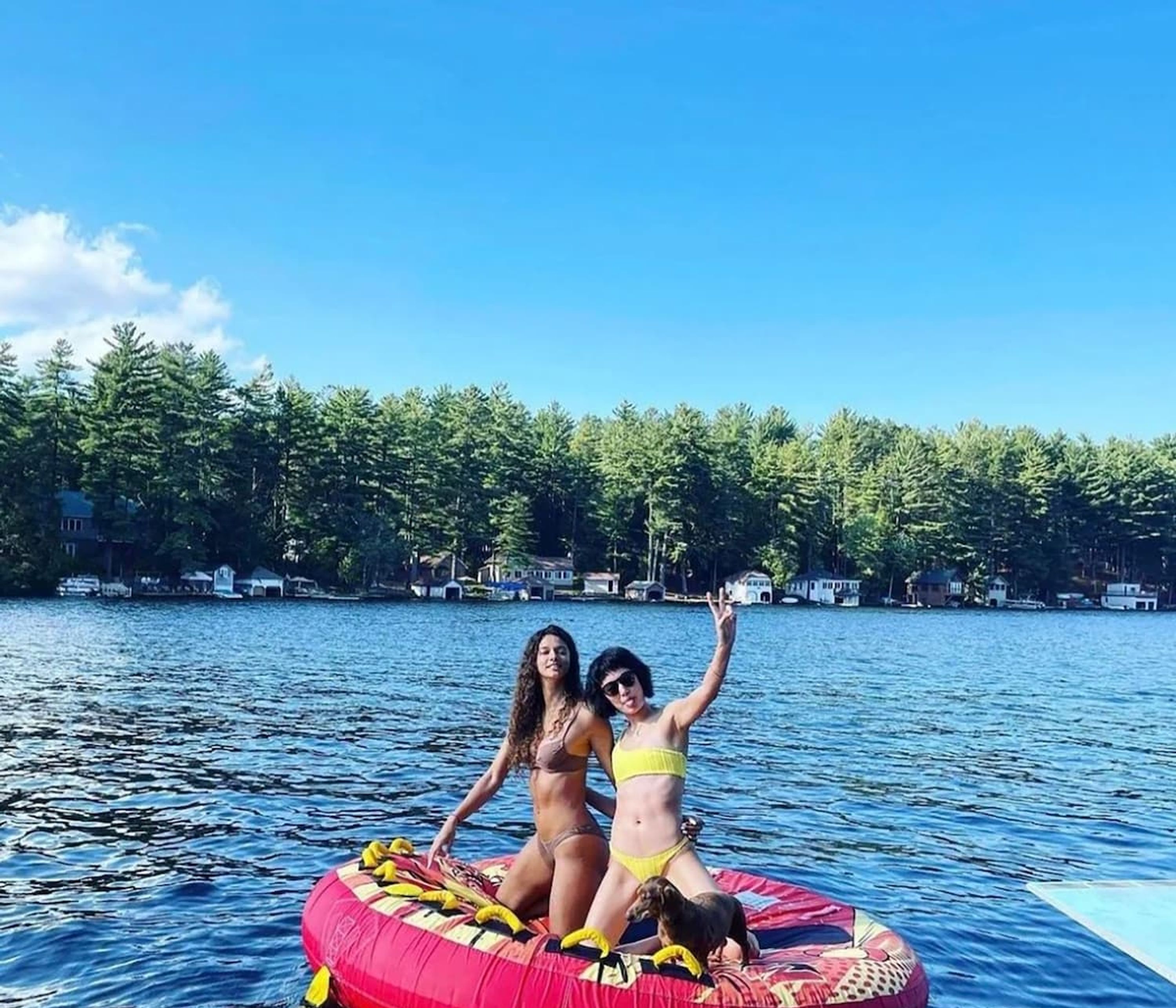 Two people on a red inflatable raft with a dog, surrounded by calm water and a forested shoreline.