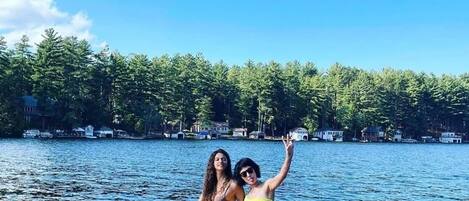 Two people on a red inflatable raft with a dog, surrounded by calm water and a forested shoreline.