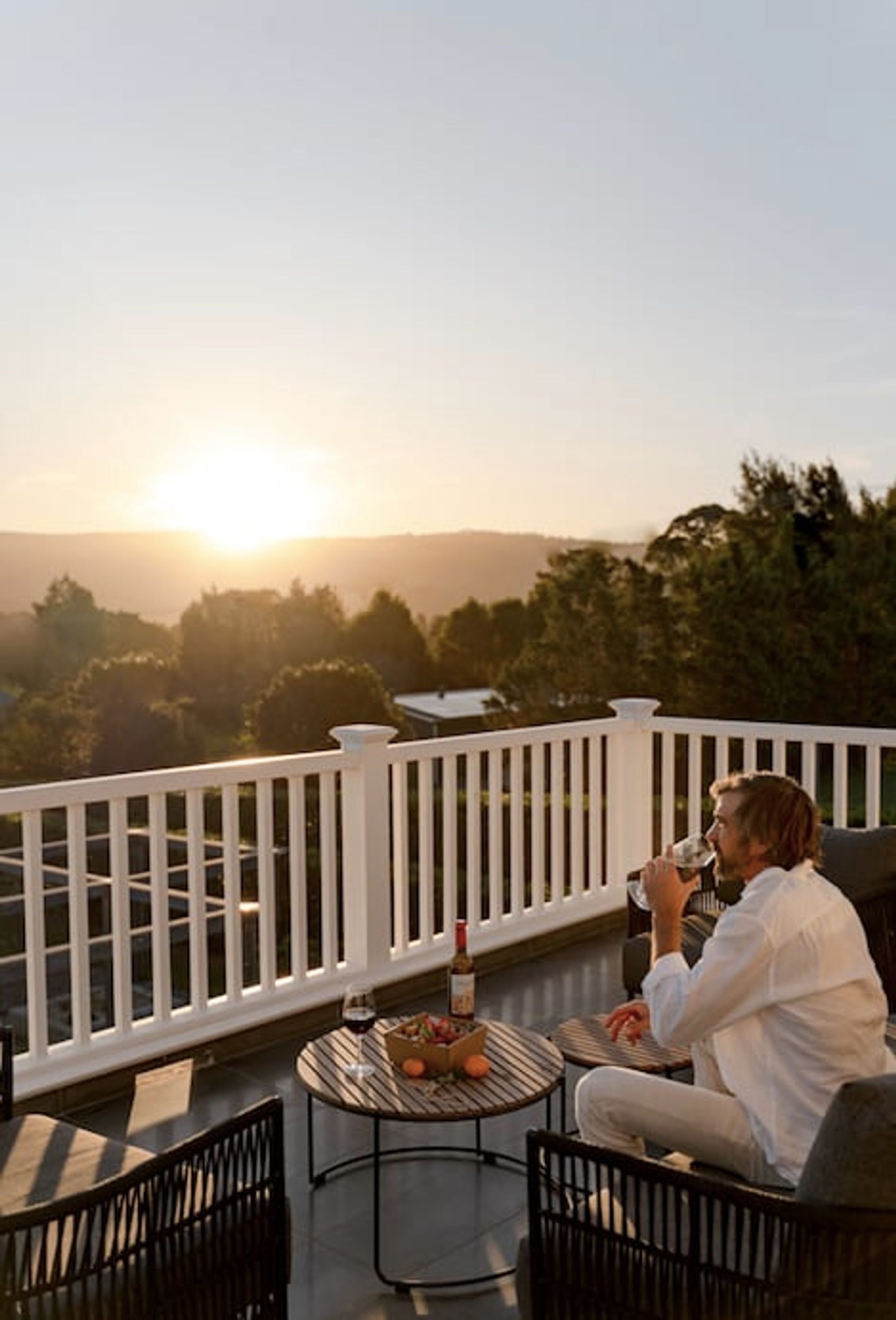 A person sitting on a balcony with a table and chairs, overlooking a scenic view of trees and a body of water during sunset.