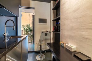 Une cuisine moderne avec un évier en acier inoxydable, un tabouret de bar en verre et une machine à café posée sur un plan de travail noir.