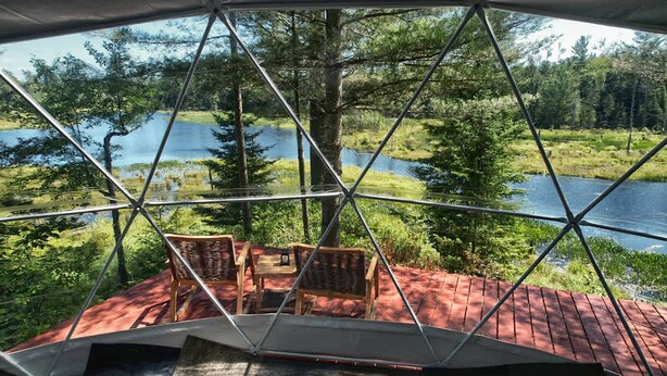 Una estructura de cúpula geodésica con techo rojo, sillas de madera y vista a un lago rodeado de árboles.