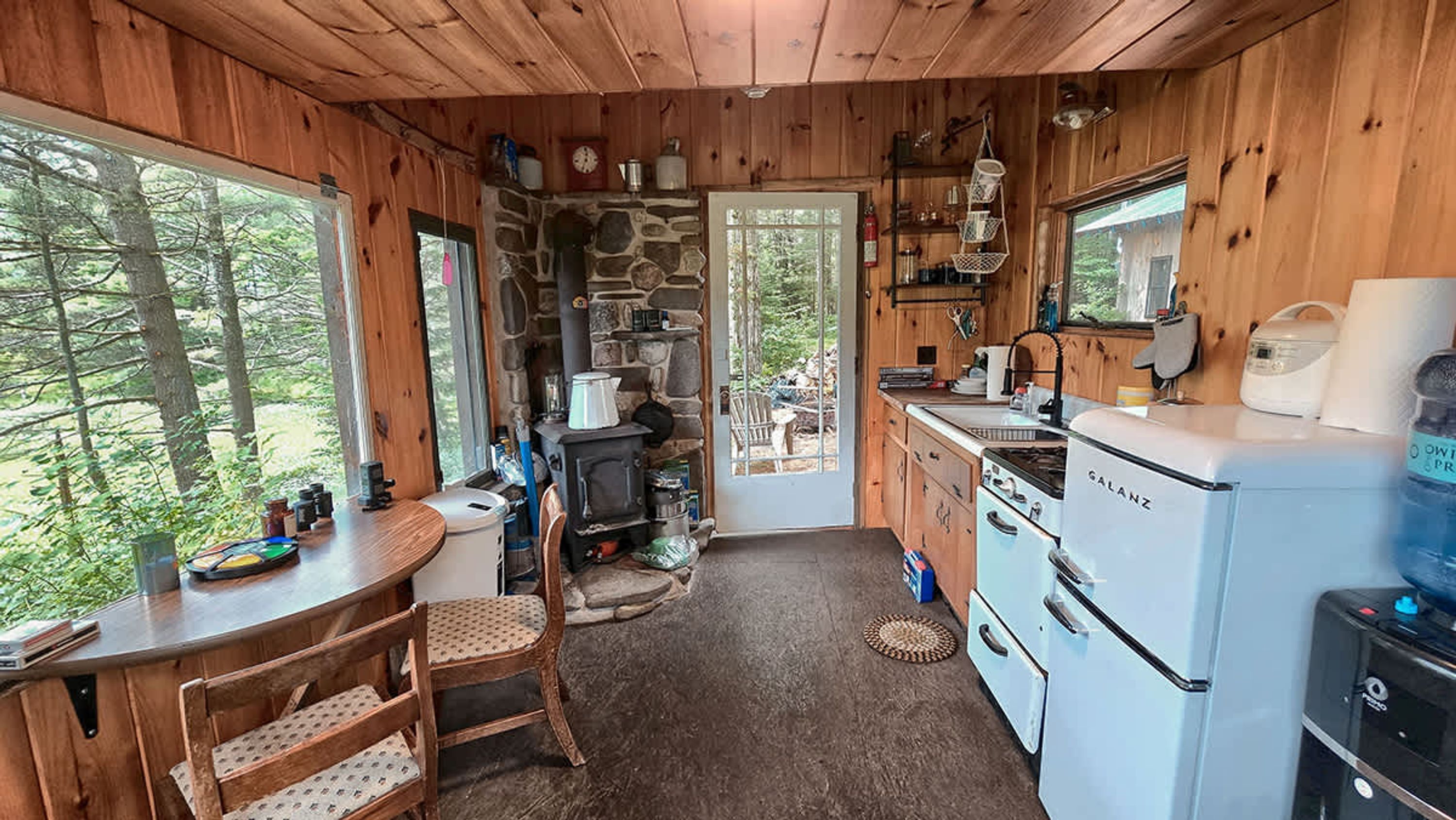 A rustic cabin kitchen with a stone wall, wooden cabinets, a sink, stove, and a window overlooking the forest.