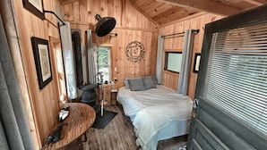 Interior de una cabaña de madera con cama, una mesita y una ventana con cortinas.