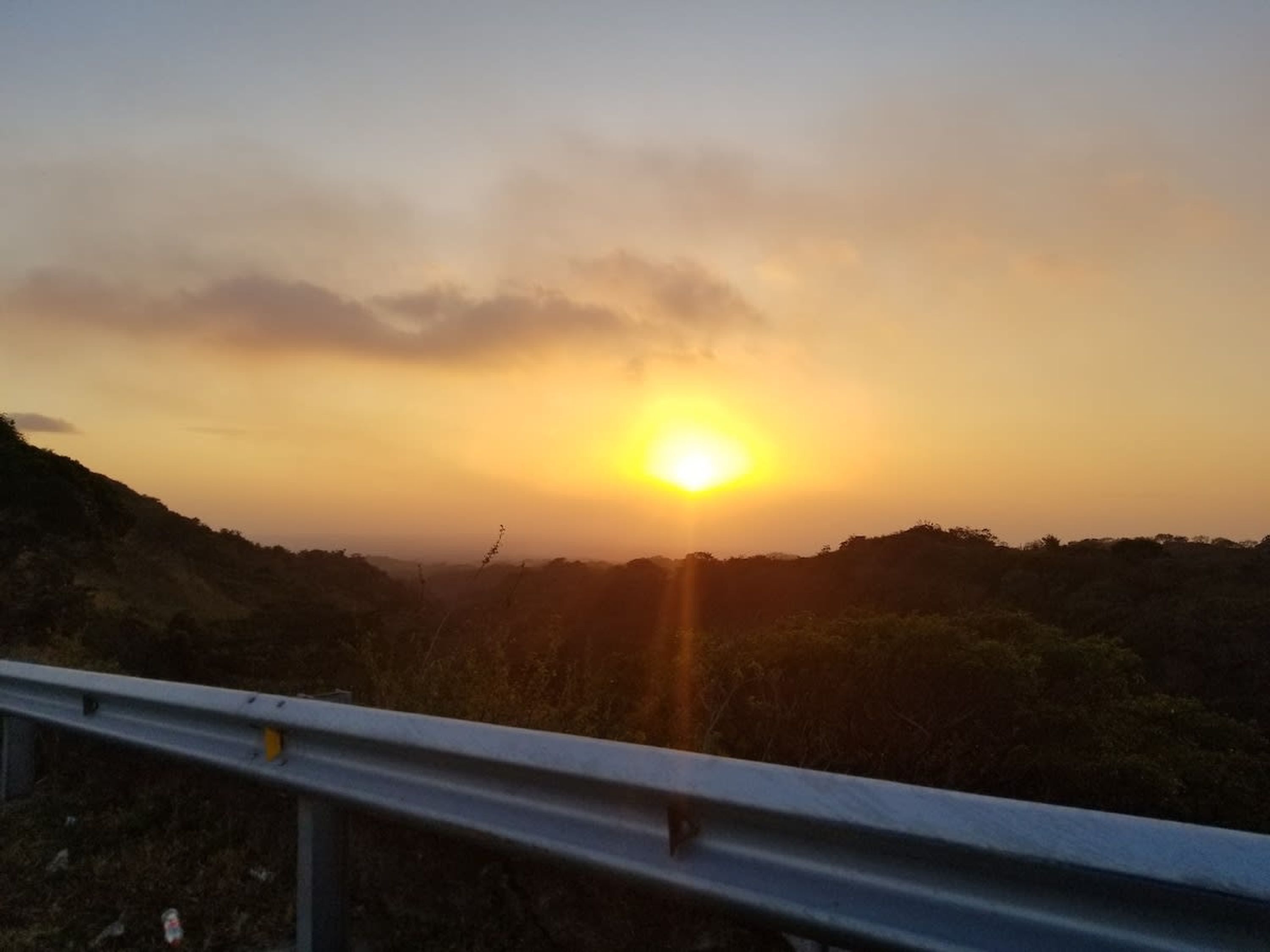 A sunset over a hilly landscape with a metal guardrail in the foreground.