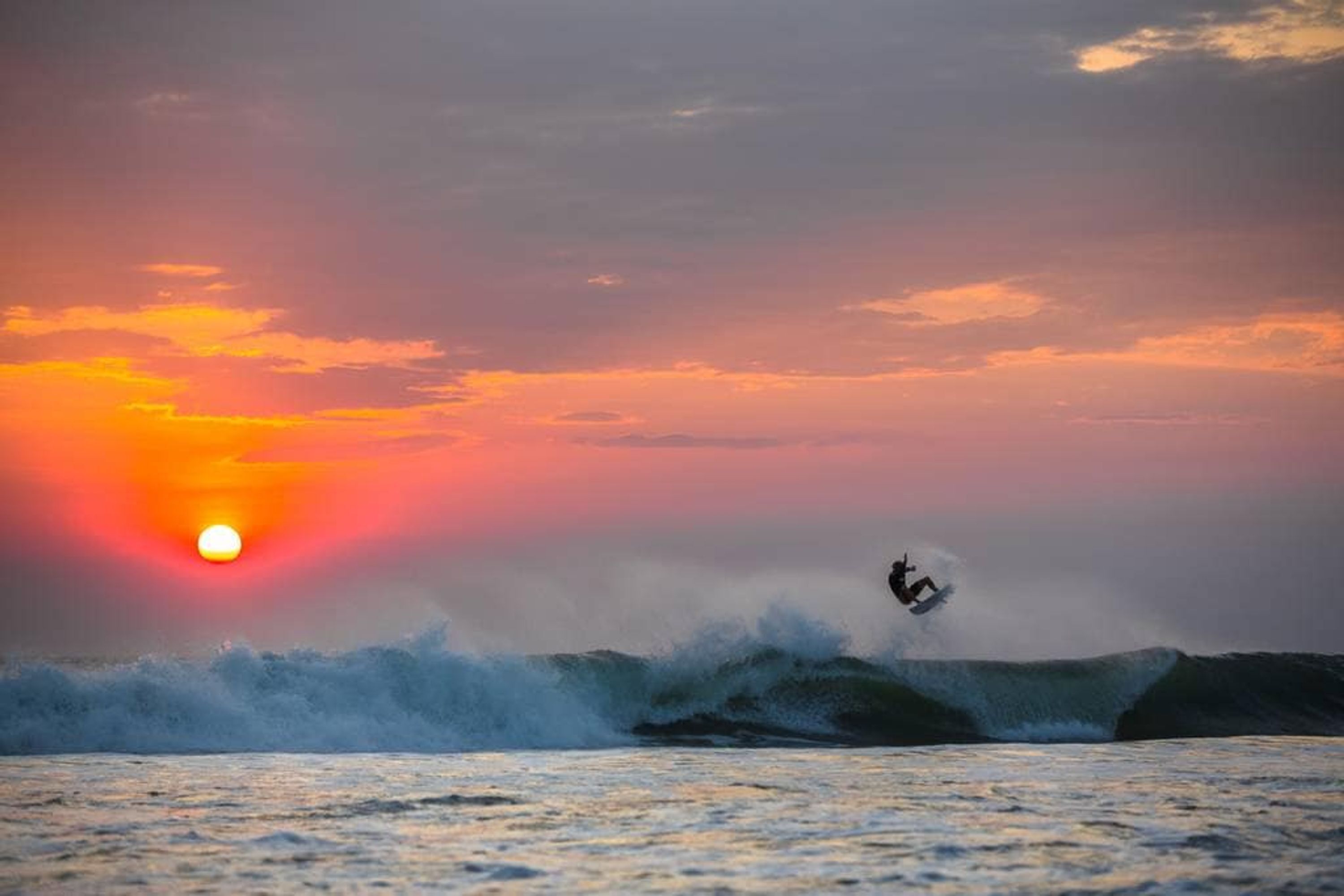 A sunset over the ocean with a surfer riding a wave.